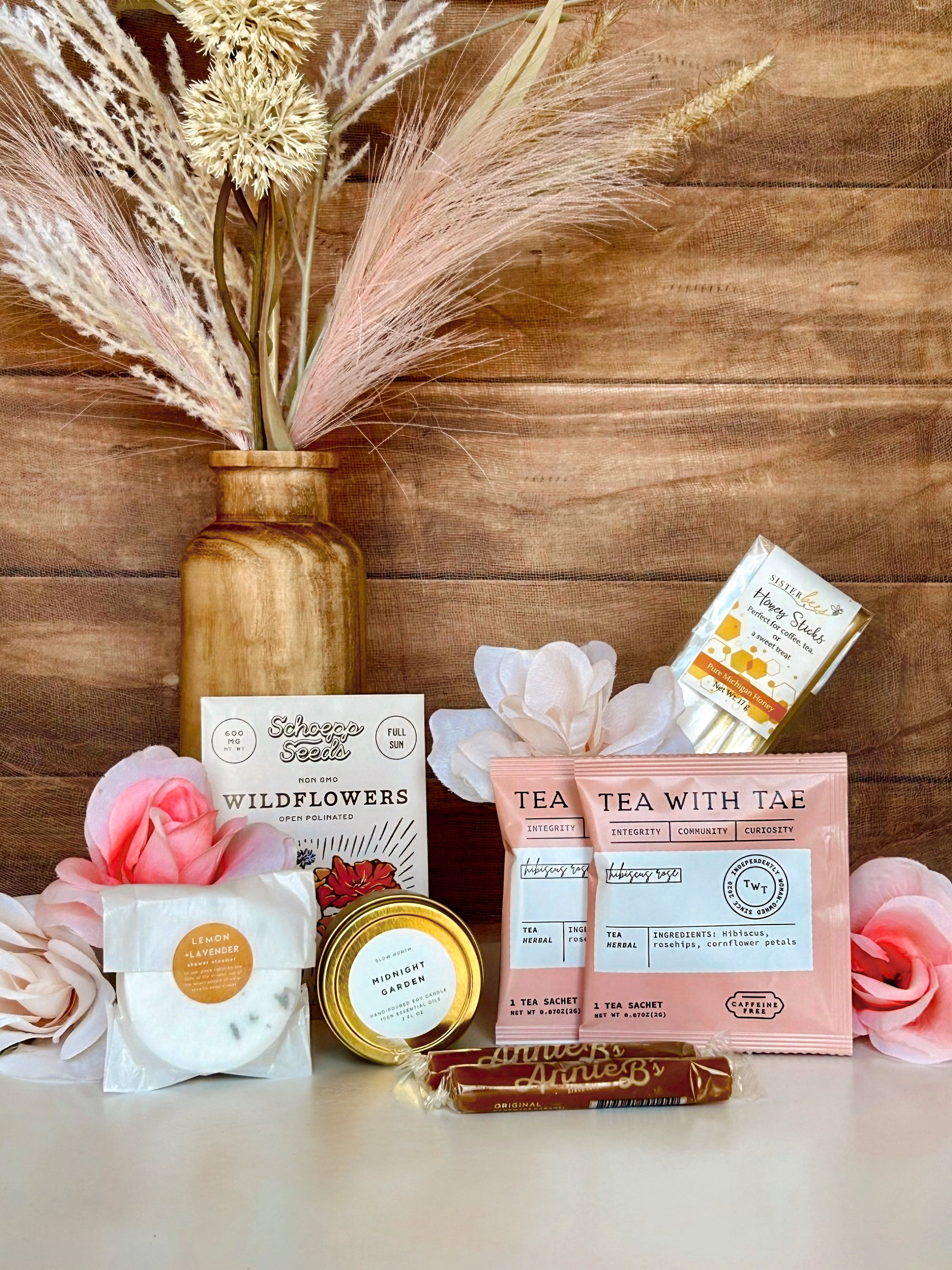 Flat lay of the Gardenlight Mini Moment gift crate, featuring floral-themed self-care items including a Midnight Garden candle, rose hibiscus tea, lemon lavender shower steamer, wildflower seeds, honey sticks, and soft caramels—styled with delicate flowers and warm, natural textures to evoke a peaceful, fairytale garden vibe.
