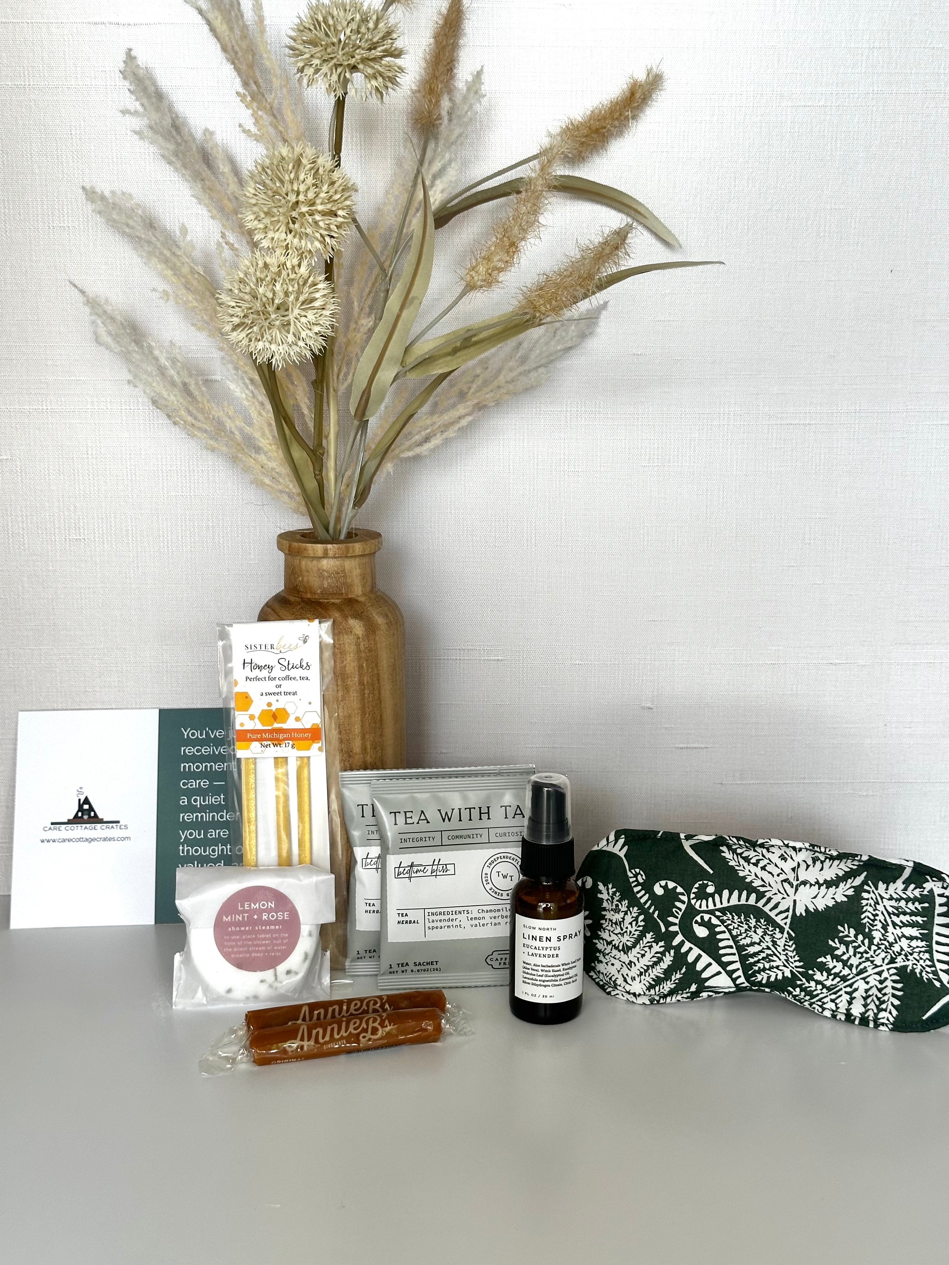 Collection of wellness products including a linen spray, tea bags, honey sticks, shower steamer, caramels, and a sleeping mask on a white surface with decorative plants.