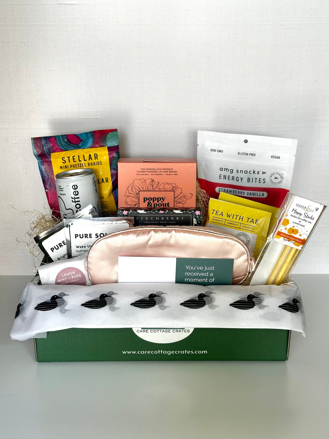 Assorted snacks and items like eye masks, sleeping mask, tea an honey, a shower steamer, cold brew and tea, honey sticks, and a poppy and pout lip duo kit, all in a box with visible branding on a white background