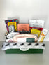 Assorted snacks and items like eye masks, sleeping mask, tea an honey, a shower steamer, cold brew and tea, honey sticks, and a poppy and pout lip duo kit, all in a box with visible branding on a white background