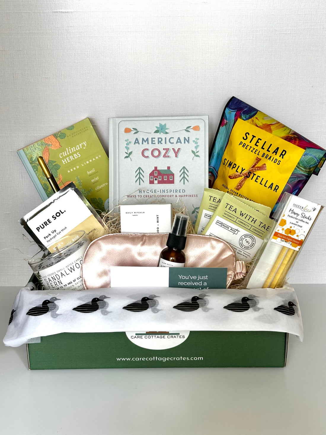 Gift box with various items including book, herbal seed kit,  tea bags, pretzels, sleeping mask, linen spray honey sticks, and and a candle on a white background.