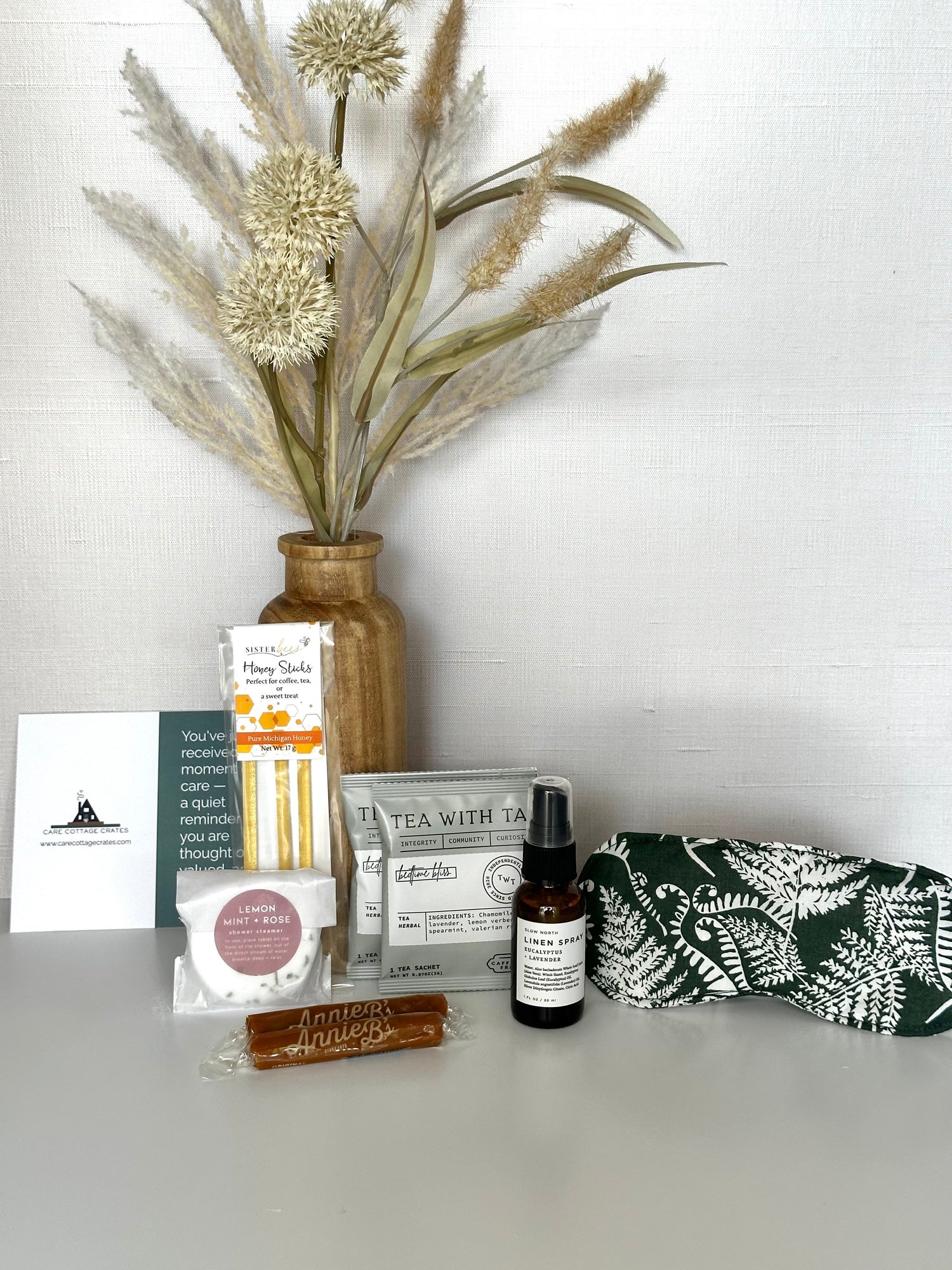 Collection of wellness products including a linen spray, tea bags, honey sticks, shower steamer, caramels, and a sleeping mask on a white surface with decorative plants.