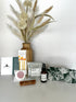 Collection of wellness products including a linen spray, tea bags, honey sticks, shower steamer, caramels, and a sleeping mask on a white surface with decorative plants.