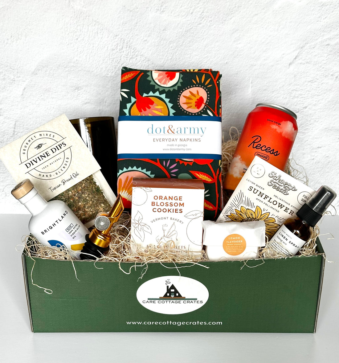 Tuscan Summer Crate from Care Cottage Crates featuring nine curated U.S.-made items including Brightland olive oil, Divine Dips Tuscan Bread Oil Mix, Dot &amp; Army napkins, orange blossom cookies, sunflower seeds, Recess mood drink, lemon lavender soap, linen spray, and an upcycled wine glass, arranged in a green gift box with natural filler.